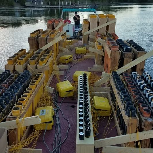 Loaded fireworks barge staged on water before a waterfront display Fireworks barge on water loaded with rows of mortar tubes, electric firing modules, and yellow shot boxes at dusk before a waterfront display