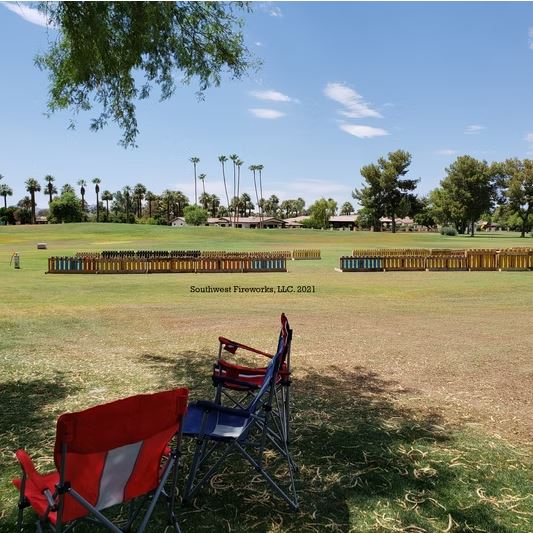 Lawn chairs facing loaded mortar racks before an Arizona daytime show setup Two empty red and blue lawn chairs facing a long line of loaded fireworks mortar racks on a green golf course with palm trees and blue sky before showtime