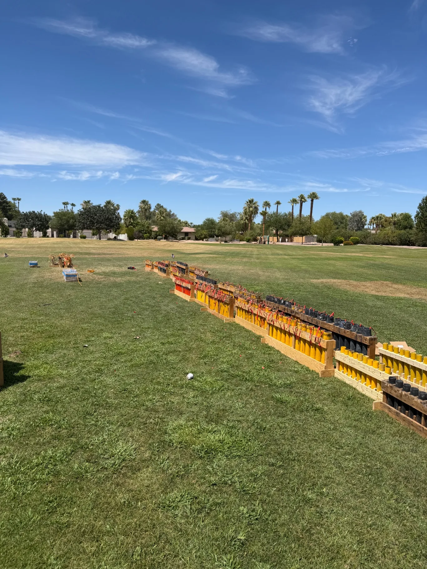 Loaded fireworks mortar racks staged on an Arizona field Row of loaded yellow and black fireworks mortar tubes secured in wooden racks on a grass field with palm trees in the background
