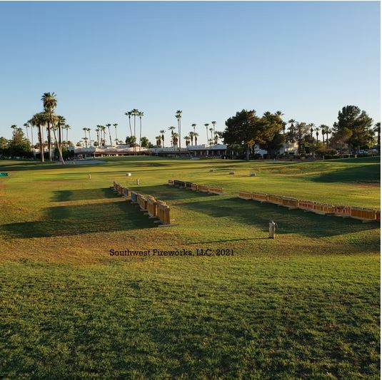 Fireworks mortar racks staged across an Arizona resort golf course Long rows of loaded fireworks mortar racks staged across a manicured Arizona golf course fairway with palm trees and a clubhouse in the background at golden hour