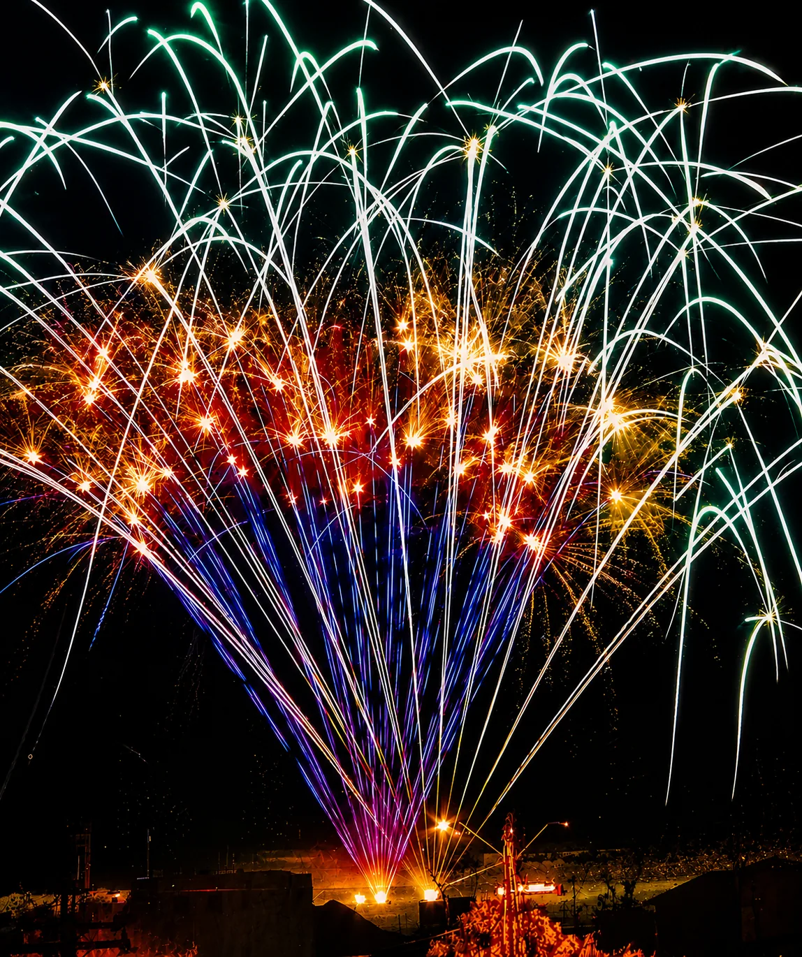 Multi-shot cake finale — green, gold, blue, and red bursts Multi-shot fireworks cake firing a rapid sequence of green, gold, blue, and red aerial bursts across the night sky